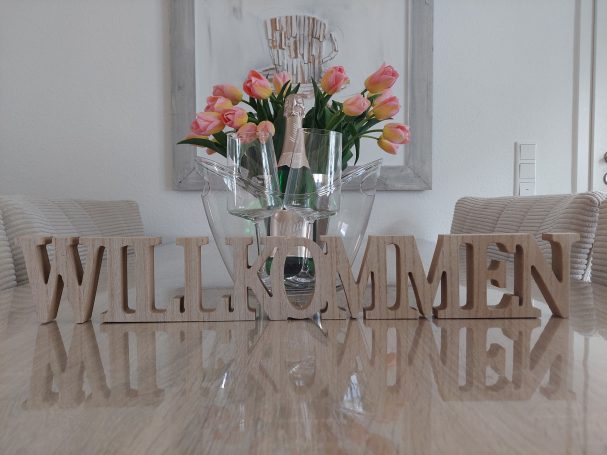 Text "WILLKOMMEN" aus Holz vor einer Blumenvase mit rosa Tulpen auf einem Tisch.